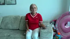 Mujer con camiseta deportiva roja sostiene un celular en un sofá.