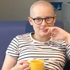 Una joven con gafas y una camiseta de rayas blancas y negras sostiene una taza amarilla y está sentada en un sofá azul. No tiene pelo.