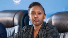 Yolande Makolo yambaye umwenda w'umukara n'umweru arimo kuvugira kuri microphone