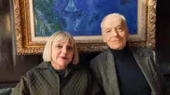 Una mujer madura con pelo claro y un hombre maduro de pelo canoso vestidos causual pero elegantemente y sentados frente a un cuadro de Chagall