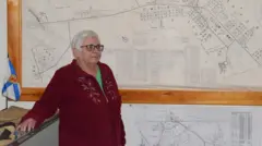 Annette Sharpe, de pé, à esquerda, olha para o outro lado, ao lado de mapas em preto e branco na parede