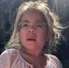 Sophia mira ligeramente fuera de cámara. Tiene el cabello largo y rubio recogido hacia atrás, y lleva unas gafas grandes de montura rosa y una blusa blanca de verano.