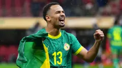 Iliman Ndiaye, visible from waist up, clenches his fists, closes his eyes and screams in delight as he celebrates winning the 2025 Africa Cup of Nations final in Rabat. He is wearing a green Senegal shirt with yellow trim and a yellow number 13 on the best and is clutching a scrunched up Senegal flag in his right hand
