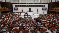 TBMM Genel Kurulu'nda oturuma katılan milletvekilleri, kırmızı Meclis koltuklarında otururken arkadan geniş bir açıyla görünüyorlar. 