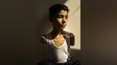 Mahmoud Ajjour, aged 9, pictured by a wall. He is wearing a white vest and has both arms missing