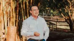 Um homem de meia-idade, Augusto Cury está ao ar livre, em um ambiente com bambus e árvores ao fundo, vestindo uma camisa social clara, cinto escuro e óculos de armação fina, enquanto sorri e mantém uma das mãos levemente levantada em gesto natural e a outra no bolso.