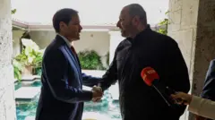 大柄で薄い口ひげをたくわえ黒いシャツ姿のウメロフ氏と、スーツ姿のルビオ氏がお互いに向き合い、笑顔で握手している。明るい中庭や池を背にしているため、逆光になっている。手前には、赤いマイクとそれを差し出す記者の右手が写っている