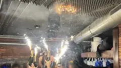 Varias personas sostenían bengalas atadas a botellas de champán en el bar de una estación de esquí suiza justo antes de que comenzara el incendio, y se puede ver una pequeña mancha naranja de fuego en lo que parece ser un techo de espuma encima de las bengalas.