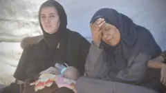 Birinin kucağında biberonla beslenen bir bebek olan iki kadın yan yana duruyor. Biri eliyle alnını hayal kırıklığı içinde tutuyor.