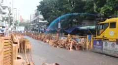 রাস্তার এক পাশে চেয়ার ভাঁজ করে রাখা এবং অন্যপাশে ভাঙা চেয়ারের স্তূপ, পুলিশের জলকামান থেকে নীল রংয়ের পানি ছোঁড়া হচ্ছে 