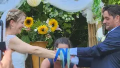 Casal sorrindo ao olhar para mãos, amarradas por fitas, no altar florido