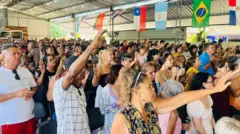 Pessoas com braços erguidos em igreja, em ambiente iluminado e com bandeiras de vários países, inclusive o Brasil