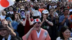 Pessoas vibram em evento ao ar livre, algumas com bandeiras símbolos do Japão; no centro, uma mulher com quimono tradicional