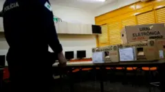 Homem fardado em frente a caixas de urna eletrônica