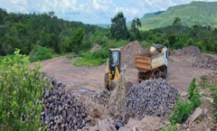 Imagem de pedreira à distância, com um caminhão e outro equipamento, com vegetação ao fundo