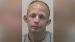 Mugshot of Adam Hall. He is clean shaven and balding with short fair hair, a drooped expression on his face and large ears.