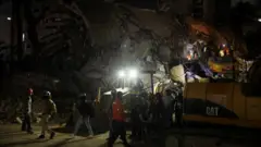 Equipo de rescate utiliza maquinaria pesada para buscar entre los escombros de un edificio colapsado, iluminado por potentes focos en plena oscuridad.