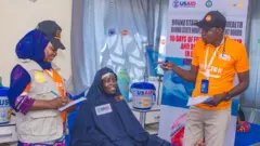 Woman wearing USAID-branded hijab on a hospital bed with two health workers