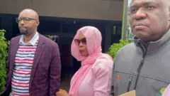 Béatrice Munyenyezi ahagaze hagati y'abagabo babiri. Yambaye ikanzu y'iroza iteye ipasi na fulari y'iroza yashyize mu mutwe no mu ijosi. Imyambaro y'iroza iranga imfungwa mu Rwanda. Yambaye n'indorerwamo. Ifoto yo ku wa gatatu, ku itariki ya 17 Ukuboza (12) mu mwaka wa 2025. 