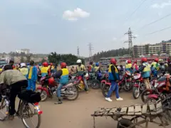 Ku gituro ca moto hari abamotari benshi bamwe bicaye ku moto zabo abandi bazihagaze iruhande bambaye casque n'udukoti tw'amabara y'umuhondo n'ubururu, imbere hari n'umuntu ari kw'igare, inyuma yiwe hari igicugutu i Muhanga, Kanama, 2025
