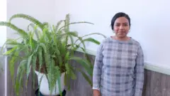 Eduarda Soares, de vestido xadrez cinza e cabelos escuros presos, sorrindo discretamente para a câmera, ao lado de uma samambaia