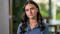 Uma mulher está sentada de frente para a câmera. Ela tem cabelo castanho comprido e pele branca bronzeada. Está usando uma camisa azul-marinho com listras brancas e sentada diante de uma janela, com a grama desfocada ao fundo