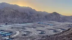 Vista de um dos empreendimentos ainda em construção na Planície de el-Raha. O sol está atrás das montanhas ao redor, enquanto o local do projeto aparece em primeiro plano, com estradas conectando os diferentes edifícios.