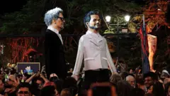 Boneco gigantes de Wagner Moura e Kleber Mendonça Filho na rua, com pessoas em volta. Elas acompanhavam cerimônia do Oscar em frente ao Cinema São Luiz, em Recife