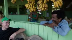 Dom sorrindo ao conversar com homem apoiado em balcão na varanda, perto de bananas