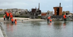Varias personas, algunas con chalecos salvavidas naranjas, se adentran en aguas poco profundas con redes junto a una orilla rocosa, donde también se ven una sauna de madera y una pequeña construcción sobre una plataforma.