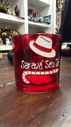 Copo transparente com a palavra “Saravá” impressa em vermelho, exposto em prateleira de loja de artigos religiosos. Ao fundo, outros objetos de culto afro-brasileiro, como imagens e velas coloridas.