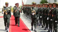 रातो कार्पेटमा नेपाली सेनाका प्रधानसेनापति अशोकराज सिग्देल, उनको दायाँबायाँ तरबार बोकेका दुई सैनिकहरू। बायाँपट्टि सतर्क मुद्रामा लामबद्ध सैनिकहरू 