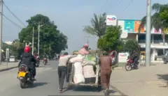 Abagabo babiri barimo gusunika imifuka irimo ibicuruzwa iri mu kinyabiziga cy'imitende, undi mugabo agitwaye. Mu muhanda hari moto ziriho abantu, hirya hari inzu ndende. 