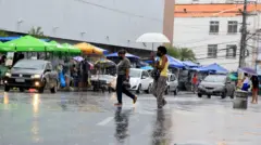chuva em rua movimentada no Brasil. Mulheres andam em faixa de pedreste, uma delas usa um guarda-chuva