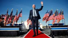 Stephen Miller chega para um evento de campanha do então candidato republicano à presidência Donald Trump, no aeroporto de Lancaster, em Lititz, na Pensilvânia, no dia 3 de novembro de 2024. Ele caminha por um tapete vermelho, vestindo terno e acenando, rodeado de bandeiras dos Estados Unidos.