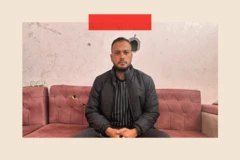 Moumen al Natour sentado en un sillón mirando a la cámara. Viste una camiseta negra con rayas verticales blancas, un abrigo y pantalones negros. Tanto el sillón como la pared detrás tienen agujeros de bala.