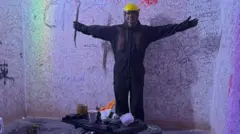 Mulher de pé com os braços abertos, com roupa de proteção, em uma sala da fúria