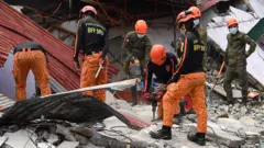 Спасатели в красных брюках и черных форменных рубашках на руинах дома. Двое из них сверлят фрагменты здания, еще один держит лопату