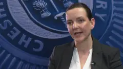 Hannah Friend, com seu cabelo escuro preso e vestindo um blazer sobre uma camisa branca, fala para a câmera com o brasão da Polícia de Cheshire visível em uma parede ao fundo