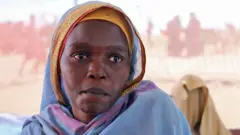 Retrato de uma mulher com um lenço azul na cabeça, mostrando apenas o busto. Ao fundo, vê-se a tela de uma tenda e algumas figuras estão desfocadas.
