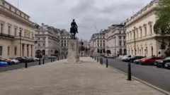 Uma estátua equestre em um pedestal de pedra fica no centro de Waterloo Place, com grandes edifícios históricos em ambos os lados da rua larga no centro de Londres.