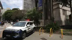 Uma viatura policial com as luzes acesas está estacionada na calçada em frente à biblioteca – um grande prédio cinza – enquanto pedestres caminham ao fundo. Um policial de braços cruzados está parado ao lado da viatura.