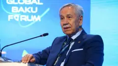 Bülent Arınç lacivert bir takım elbise giyip bir konuşma yapıyor. Arkasındaki mavi duvarda "Global Bakü Forum" yazısı okunuyor
