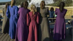 Mulheres com roupas longas e penteados de costas, em área externa e ensolarada