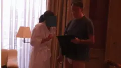 Mandelson aparece ao lado de uma mulher vestida com um robe branco. O rosto dela está oculto. Ele veste uma camiseta azul e cueca branca. Ele segura o que parece ser um iPad.