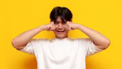 Um homem esfrega os olhos enquanto chora de forma dramática, diante de um fundo amarelo vibrante