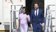 A man and woman in lilac walk down steps from a plane