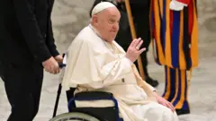 papa Francisco sendo empurado em sua cadeira de rodas. Ele sorri sutilmente e acena