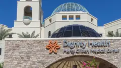 Будівля медичного центру Dignity Health Mercy Gilbert Medical Center. На передньому плані видно кам’яну стіну з назвою лікарні, позаду — світла будівля з великими скляними куполами й пальмами навколо.