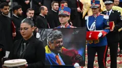 Lucescu'nun cenaze töreninde fotoğrafı ve nişanı askerler tarafından taşınıyor. Kadın askerler, gül yaprakları dökülmüş merdivenlerden aşağı yürürken etraflarında din adamları görülüyor